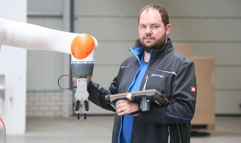  Auf dem Bild ist ein Mann in einer Lagerhalle zu sehen, der eine Steuerungseinheit hält und damit einen Roboterarm bedient.