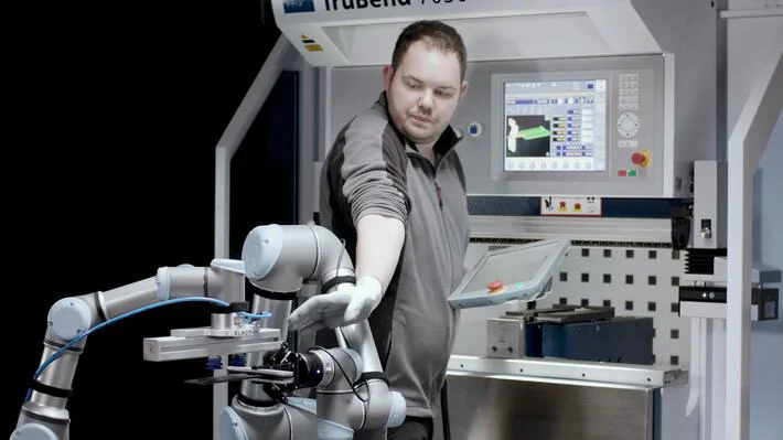 Auf dem Bild ist ein Mann in Arbeitskleidung zu sehen, der neben einem kollaborativen Roboterarm steht und eine Maschine mit einem Touchscreen-Bedienfeld bedient.