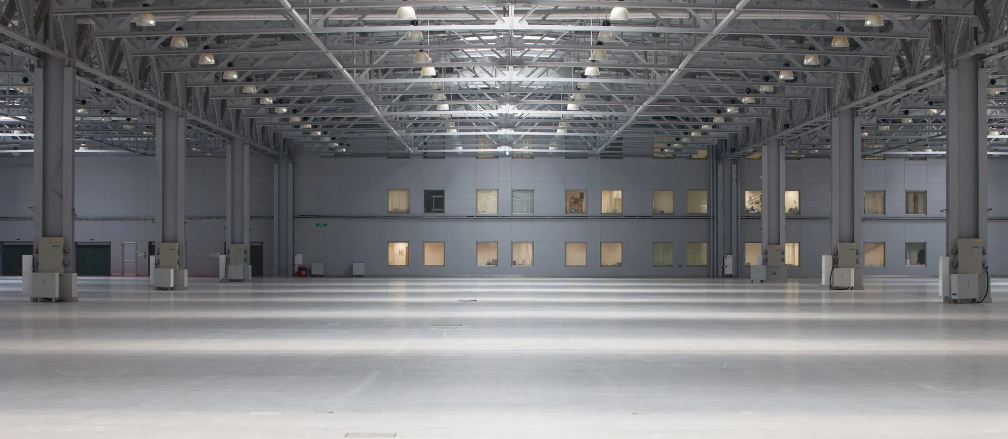Das Bild zeigt eine große, leere Industriehalle mit einer hohen Decke, ausgestattet mit Beleuchtung und einigen Schaltkästen oder Maschinen verteilt am Boden, vor einem Hintergrund mit verglasten Büroflächen.