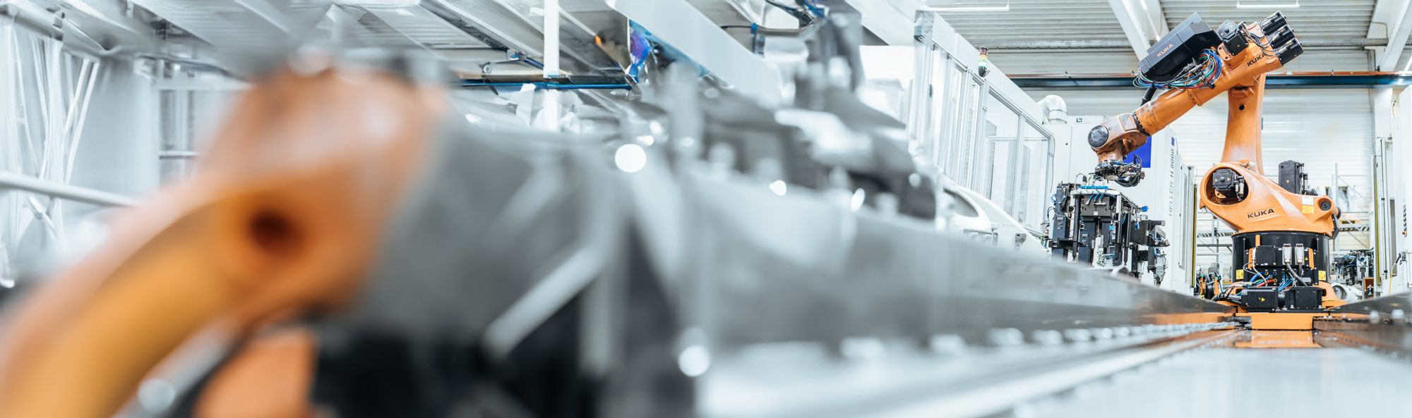 Das Bild zeigt eine hochmoderne Fertigungslinie mit einem orangefarbenen KUKA Industrieroboter, der automatisierte Aufgaben in einer sauberen und gut ausgeleuchteten Fabrikhalle ausführen.