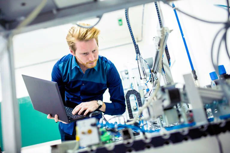 Das Bild zeigt einen konzentrierten Mann in einer Werkstattumgebung, der ein Laptop benutzt, mit einem Roboterarm oder einer Maschine im Hintergrund.