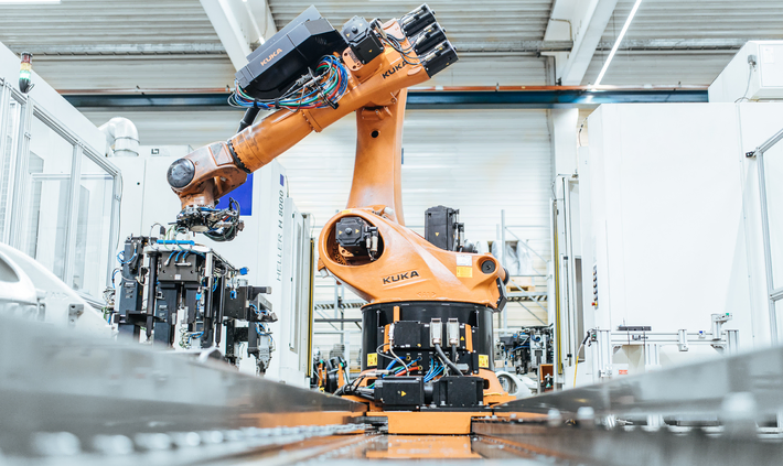 Das Foto zeigt einen großen Industrieroboter der Marke KUKA im Vordergrund, der über einer Fertigungsstraße in einer hell beleuchteten Fabrikhalle montiert ist, mit komplexen Kabeln und Komponenten, die an seinem Arm sichtbar sind. Im Hintergrund sind weitere industrielle Maschinen und Anlagen zu erkennen.