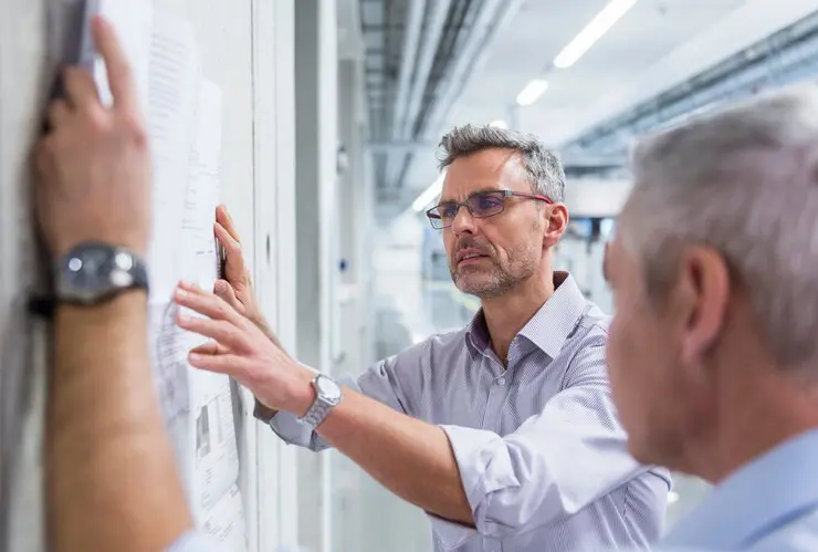 Das Bild zeigt zwei Personen die auf ein Whiteboard schreiben und dabei eine Diskussion oder Planung zu führen scheinen.