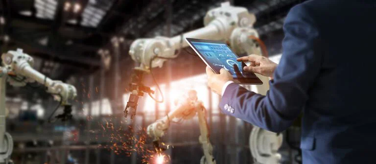 Das Bild zeigt eine Person, die ein Tablet benutzt und zwei Industrieroboter überwacht, die eine Aufgabe ausführen, bei der Funken sichtbar sind, was auf Schweißen oder eine andere Form der Materialbearbeitung hinweist.