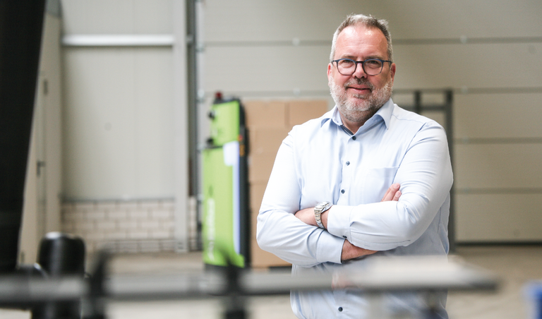 Auf dem Foto steht ein Mann mit Brille im Vordergrund, der ein hellblaues Hemd und Jeans trägt. Hinter ihm, leicht unscharf sind Teile von zwei grünen Agilox Transportrobotern zu erkennen.