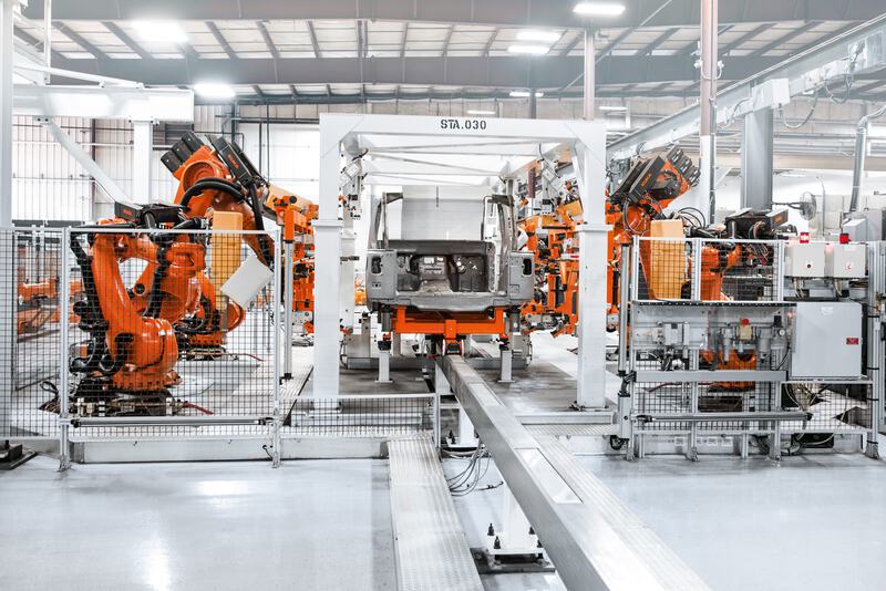 Das Bild zeigt eine hochmoderne Fertigungslinie mit orangefarbenen Industrierobotern, die um eine zentrale Vorrichtung gruppiert sind, in einem Prozess der Automobilherstellung.