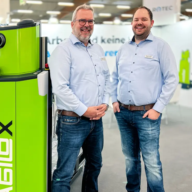 Auf dem Bild sind zwei lächelnde Männer in hellblauen Hemden und Jeans zu sehen, die vor einem grünen Agilox Transportroboter stehen.