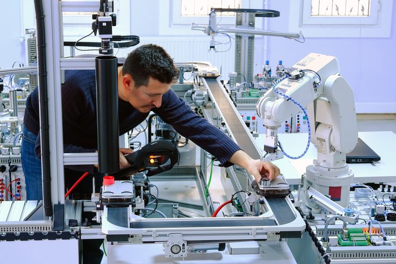 Das Bild zeigt einen Mann, der konzentriert an einer technischen Arbeitseinheit mit diversen Maschinenteilen und einem Roboterarm arbeitet, vermutlich in einer Industrie- oder Forschungsumgebung.